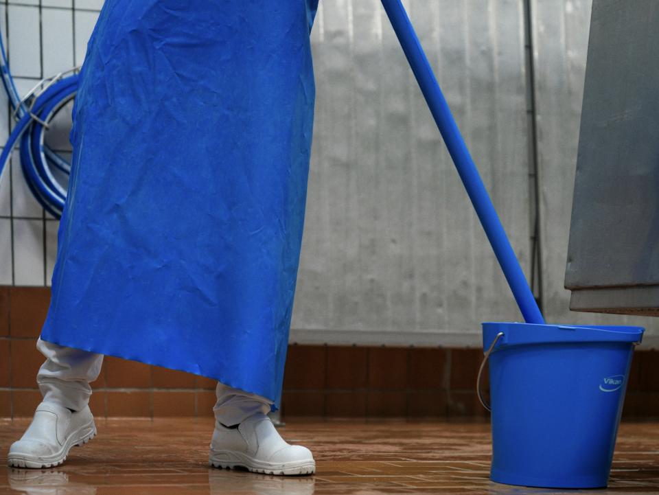 Person iført blåt forklæde og hvide sikkerhedssko, som gør rent i produktionsvirksomhed